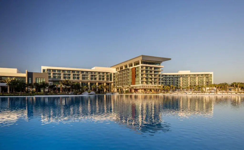Large modern resort-style building with multiple balconies reflecting on calm water at sunset.