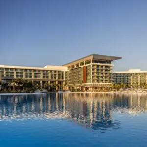 Large Modern Resort-style Building With Multiple Balconies Reflecting On Calm Water At Sunset.