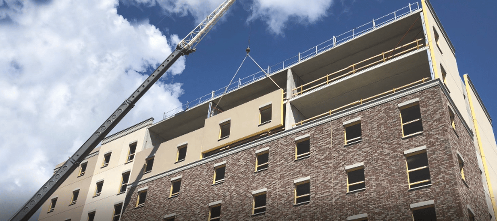 Construction site photo showing a crane lifting and installing a prefabricated exterior wall panel on a multi-story building.