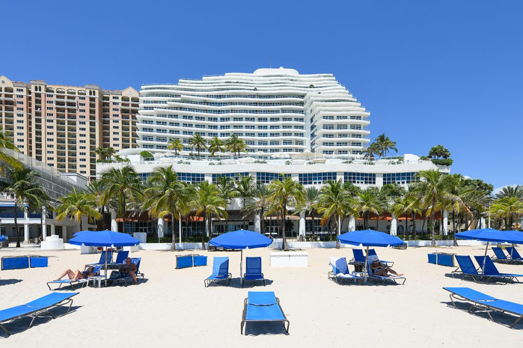 Ritz-Carlton Fort Lauderdale beachfront hotel with white facade, palm trees, and blue umbrellas in a coastal environment