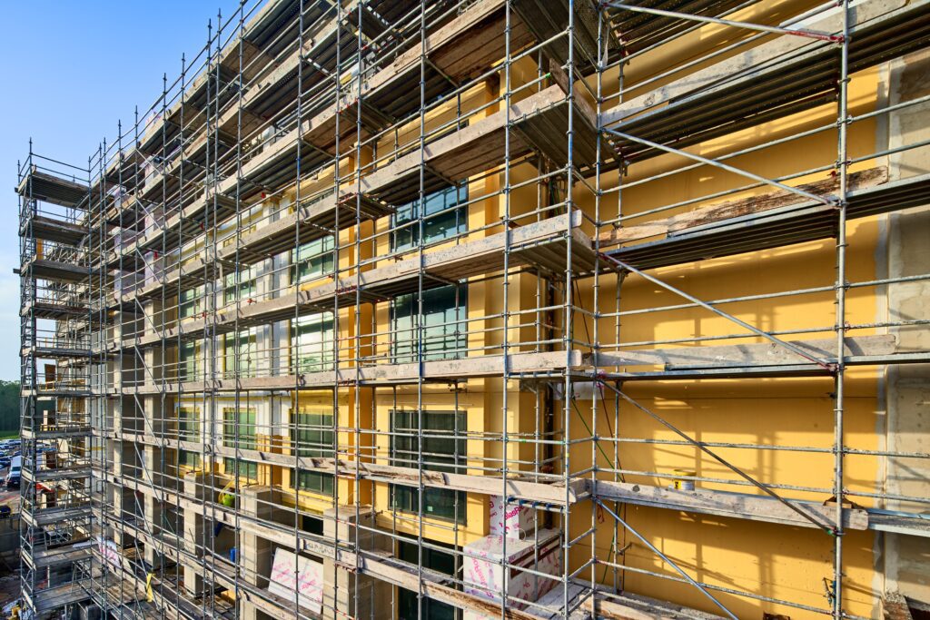 Multi-story building under construction with scaffolding surrounding exterior walls covered in yellow sheathing.