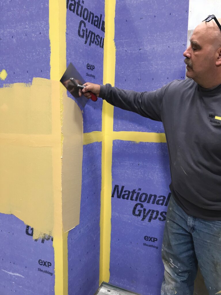 Installer using a trowel to apply a fluid-applied air barrier over gypsum sheathing with treated seams and an inside corner