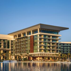Completed Multistory Hotel With A Cantilevered Roof, Glass Balconies And A Vertical Wood-look Accent Panel Overlooking A Lagoon At Sunset